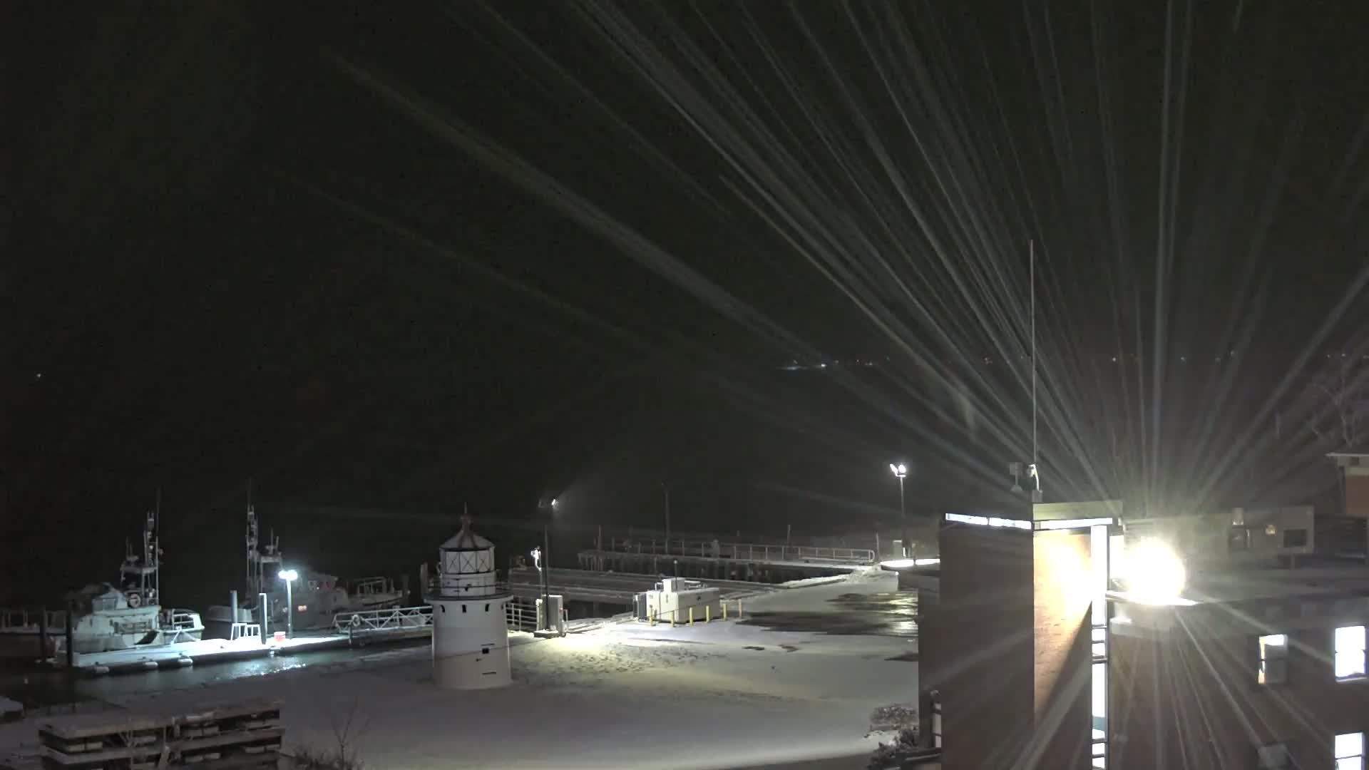 A dark, snowy night scene depicts a white lighthouse and a docked boat by a pier, with bright artificial lights creating prominent streaks as snow falls and covers the ground.