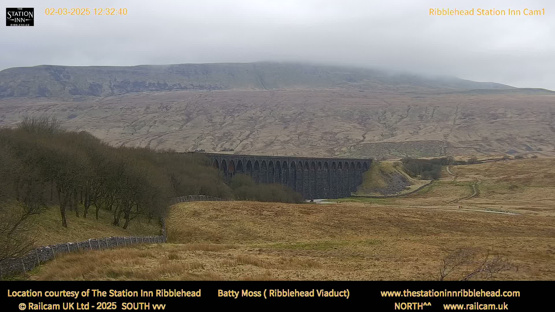 A large stone viaduct sits in a brown, grassy valley beneath a hazy, overcast mountain.