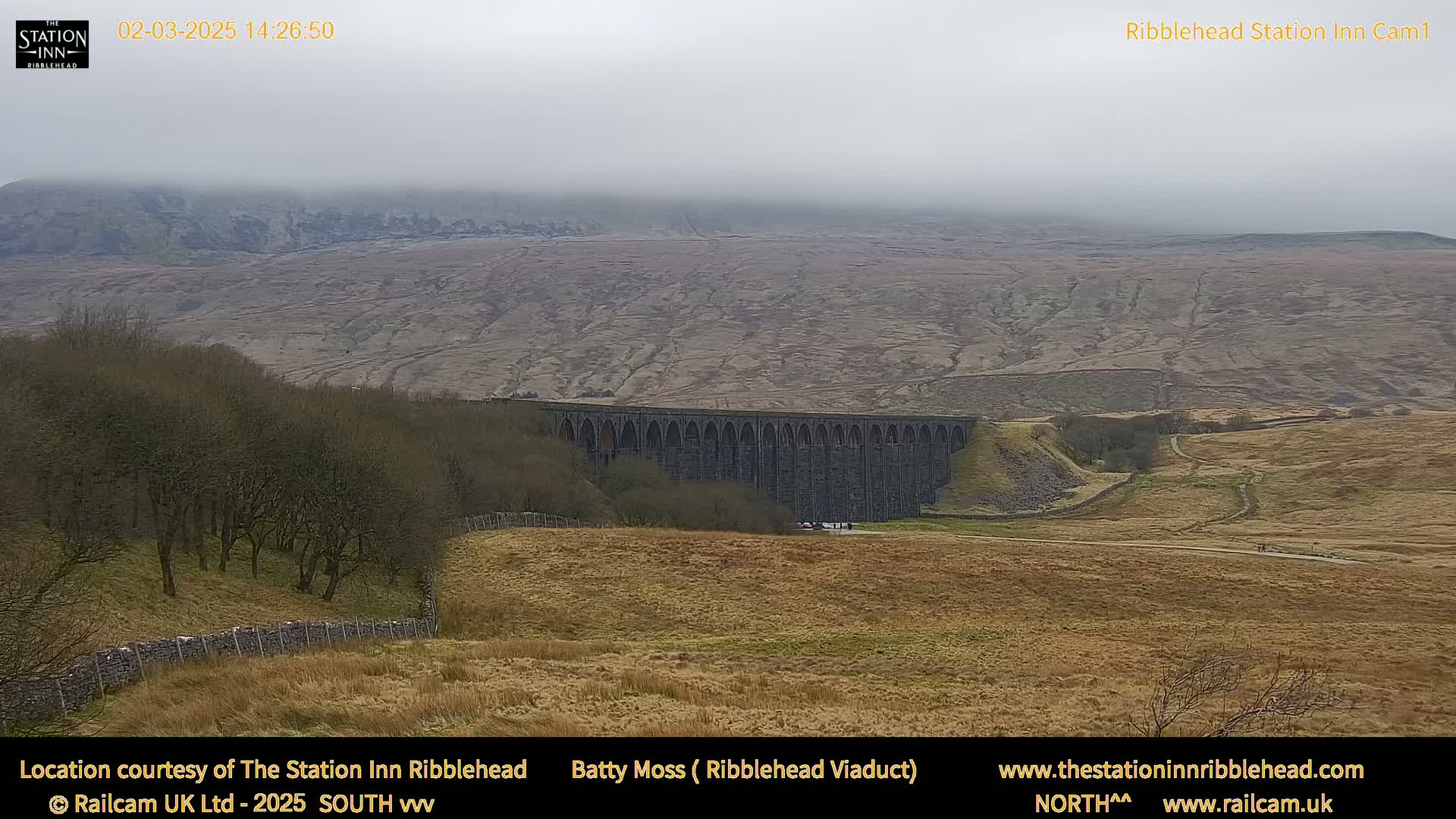 A large stone viaduct sits in a valley under an overcast sky, with dry, brown fields and distant hills.