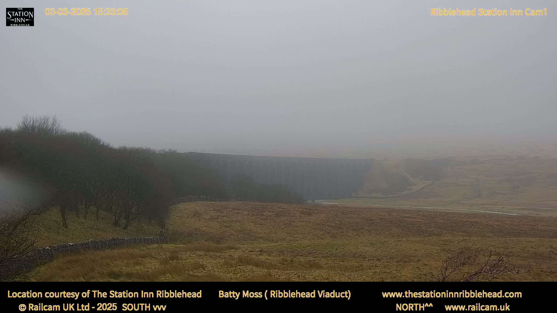 A long stone viaduct is partially visible through a dense fog over a brown, grassy landscape.