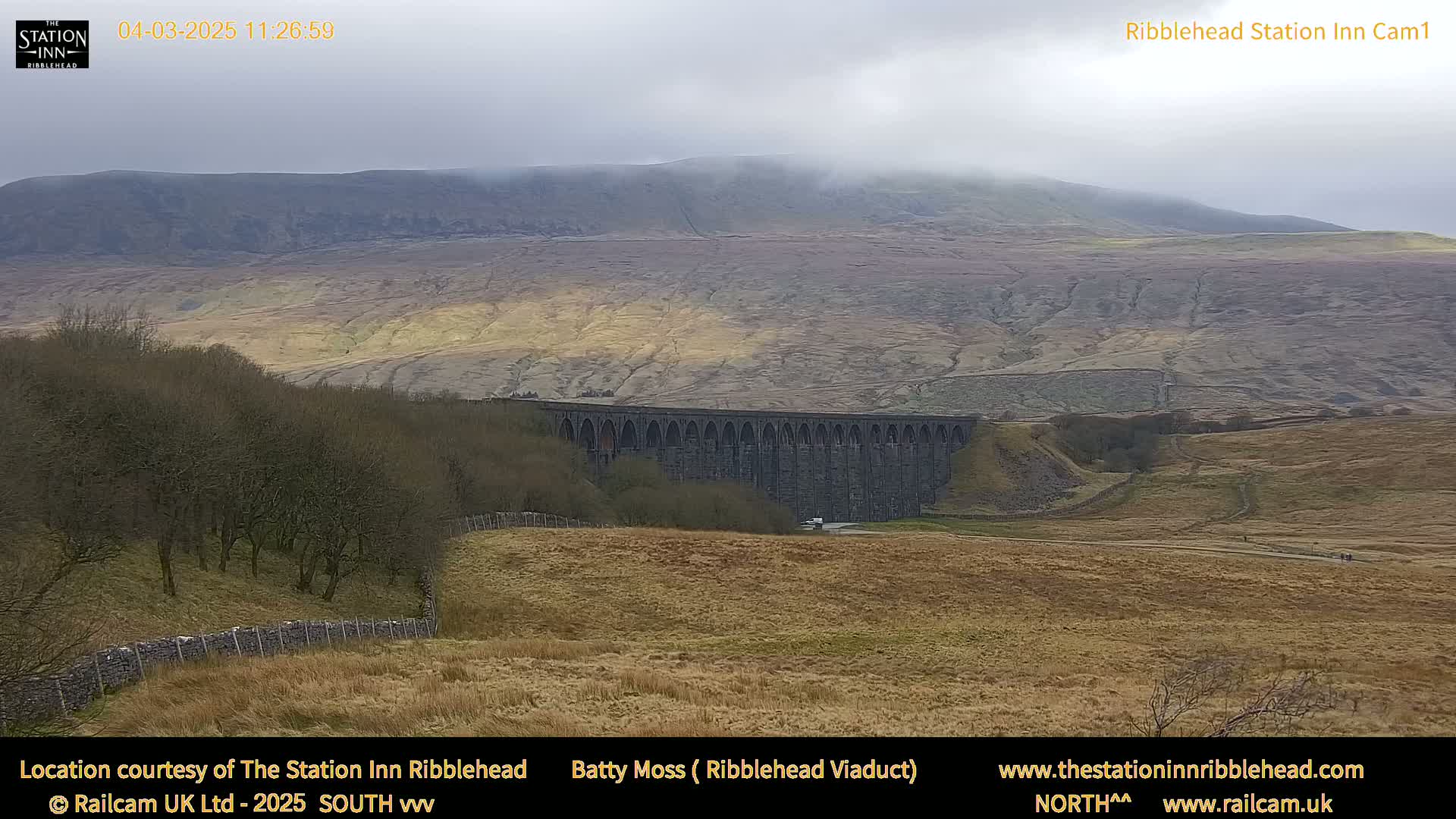 A long stone viaduct stretches across a valley, with a hazy mountain in the background under a cloudy sky.