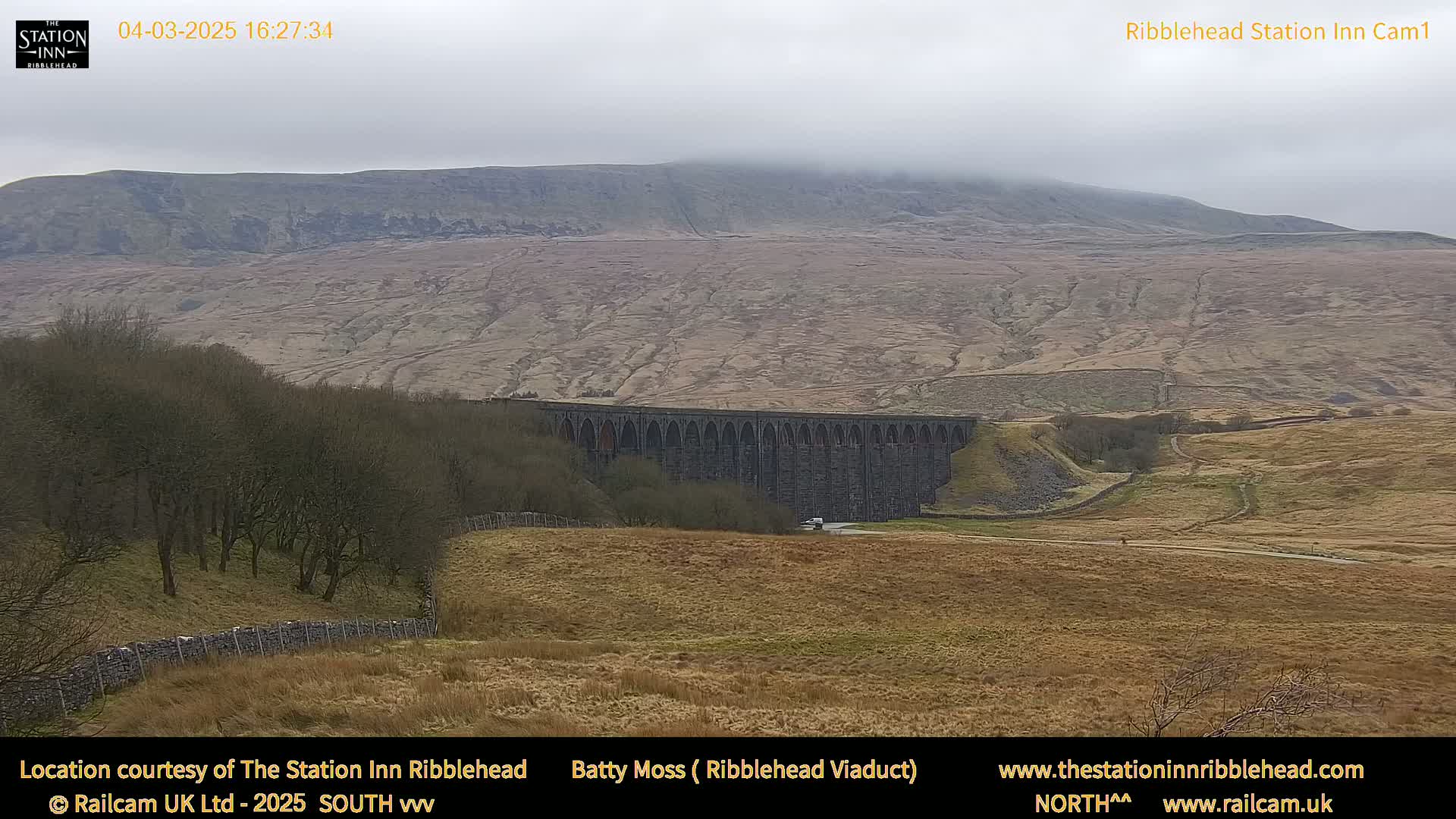 A large stone viaduct stretches across a valley under a foggy sky, with a mountain in the background and dry, brown fields in the foreground.