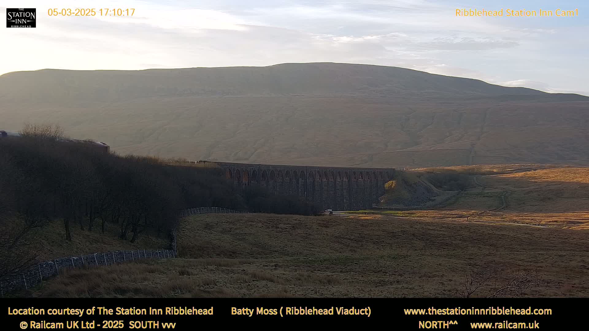 A long stone viaduct stretches across a brown, hilly landscape under a mostly clear, late afternoon sky.