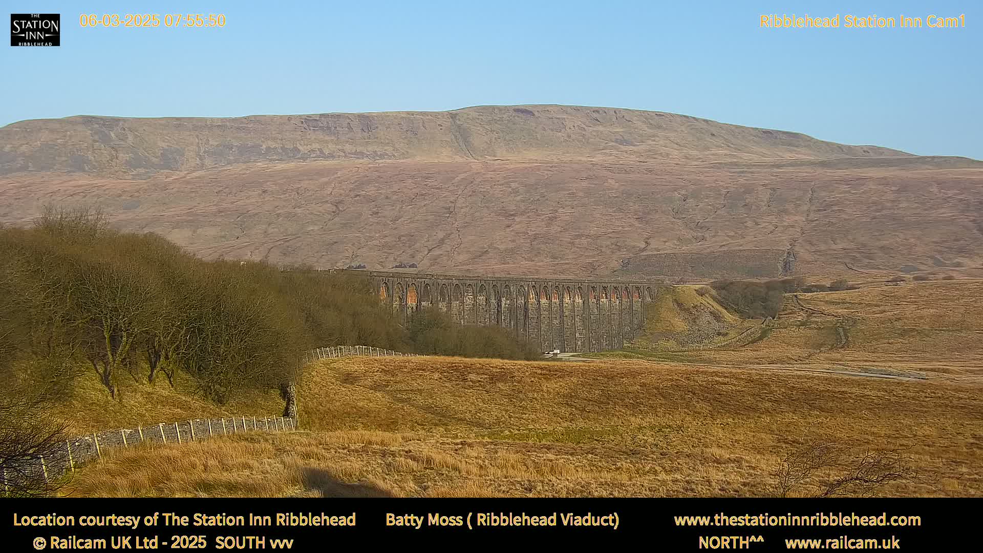 A long stone viaduct sits in a dry, brown field beneath a large, brown hill under a clear blue sky.