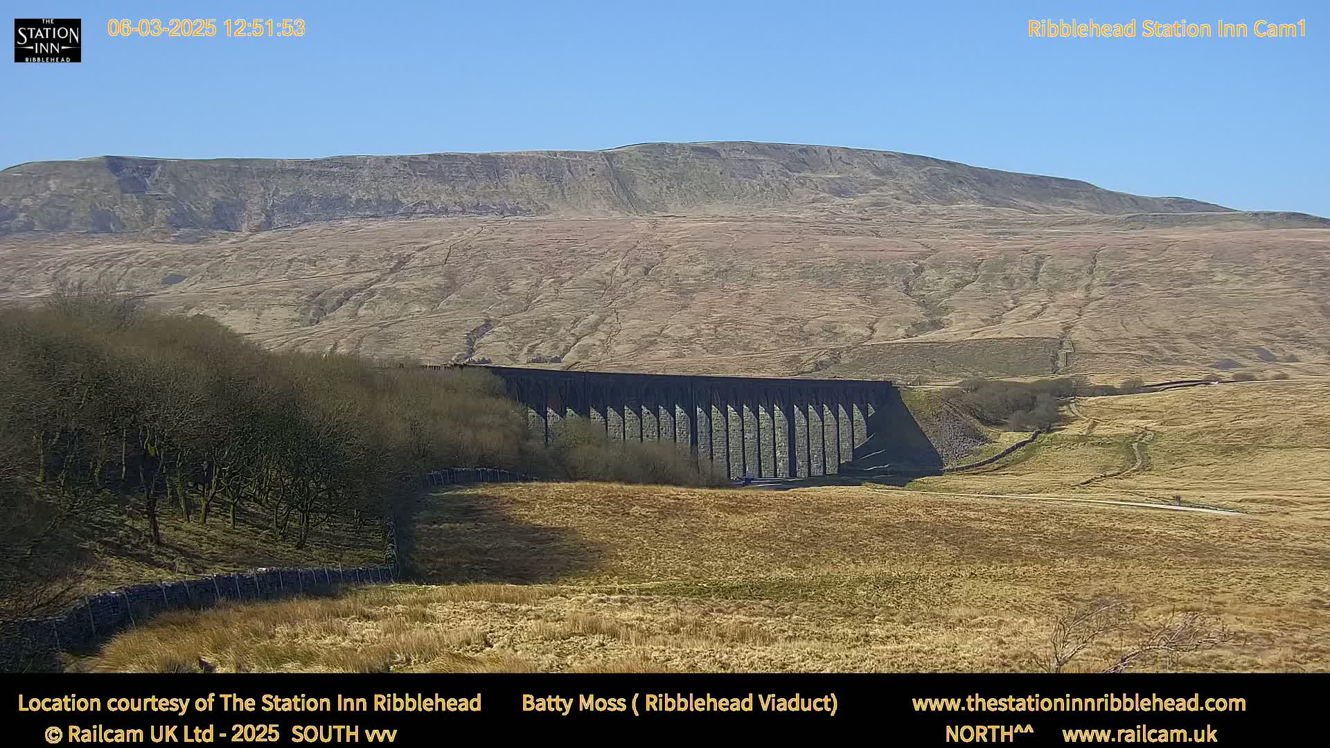 A long stone viaduct sits in a dry, brown field beneath a large, tan-colored hill under a clear blue sky.