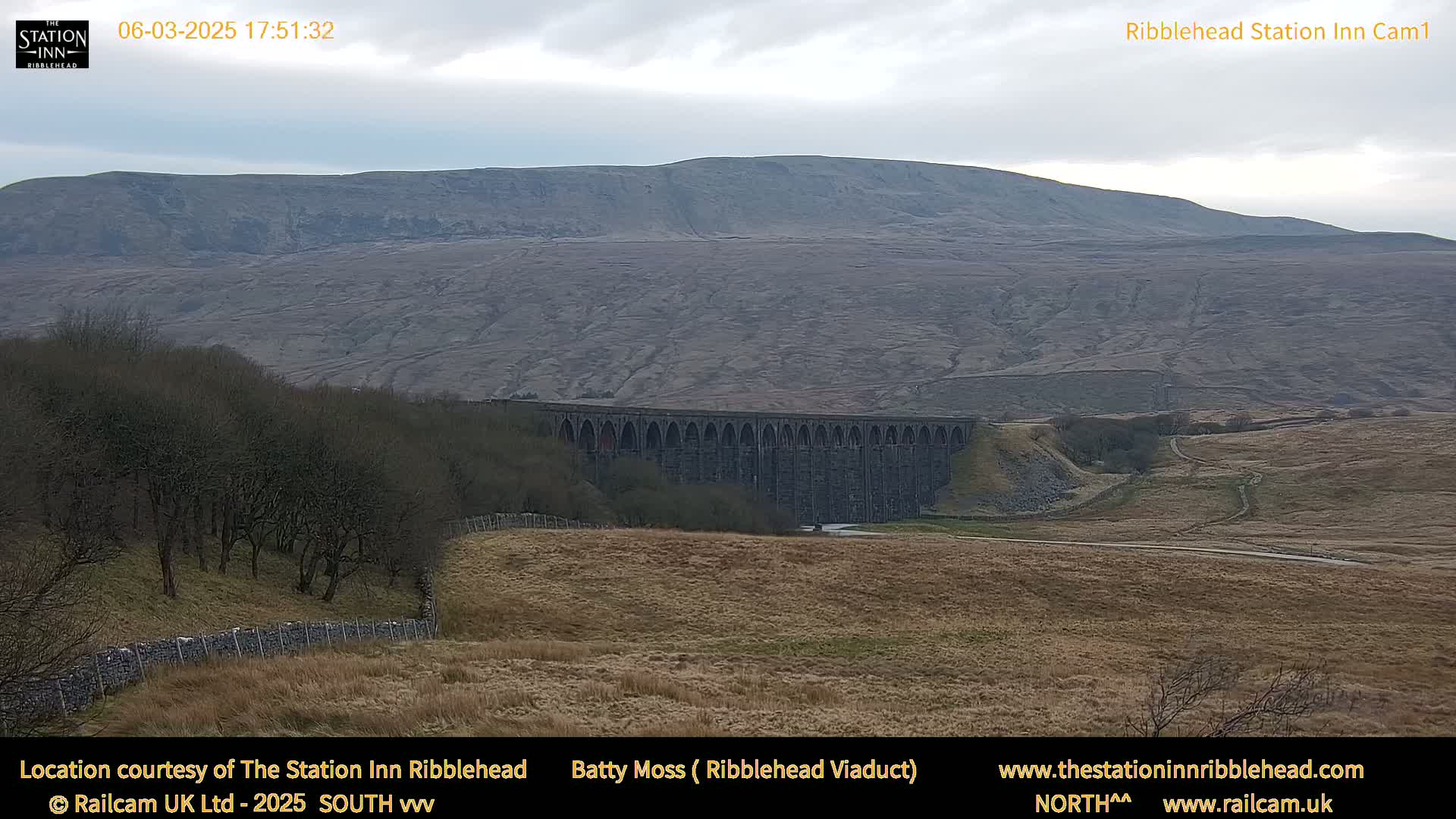 A large stone viaduct sits in a valley below a large, barren hill under an overcast sky.