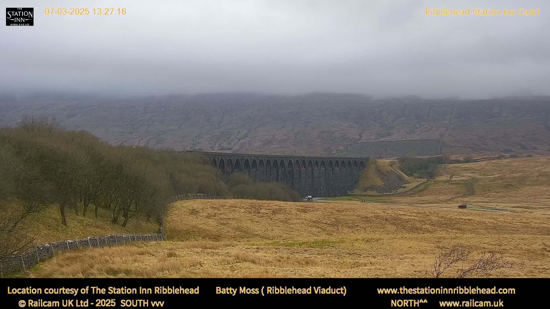 A long stone viaduct stretches across a valley under a foggy sky, with dry, tan-colored fields in the foreground and hills in the background.