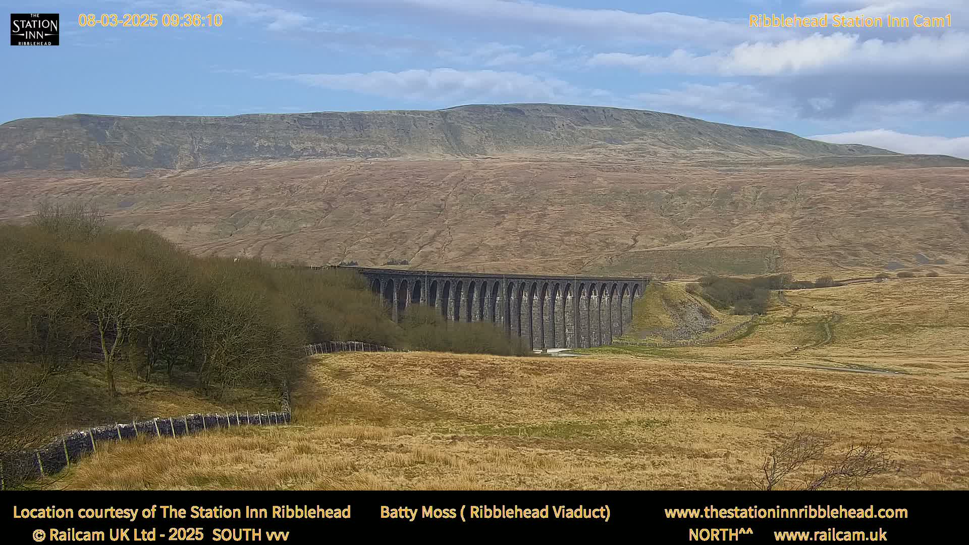 A long stone viaduct stretches across a valley beneath a large, brown hill under a mostly clear sky.