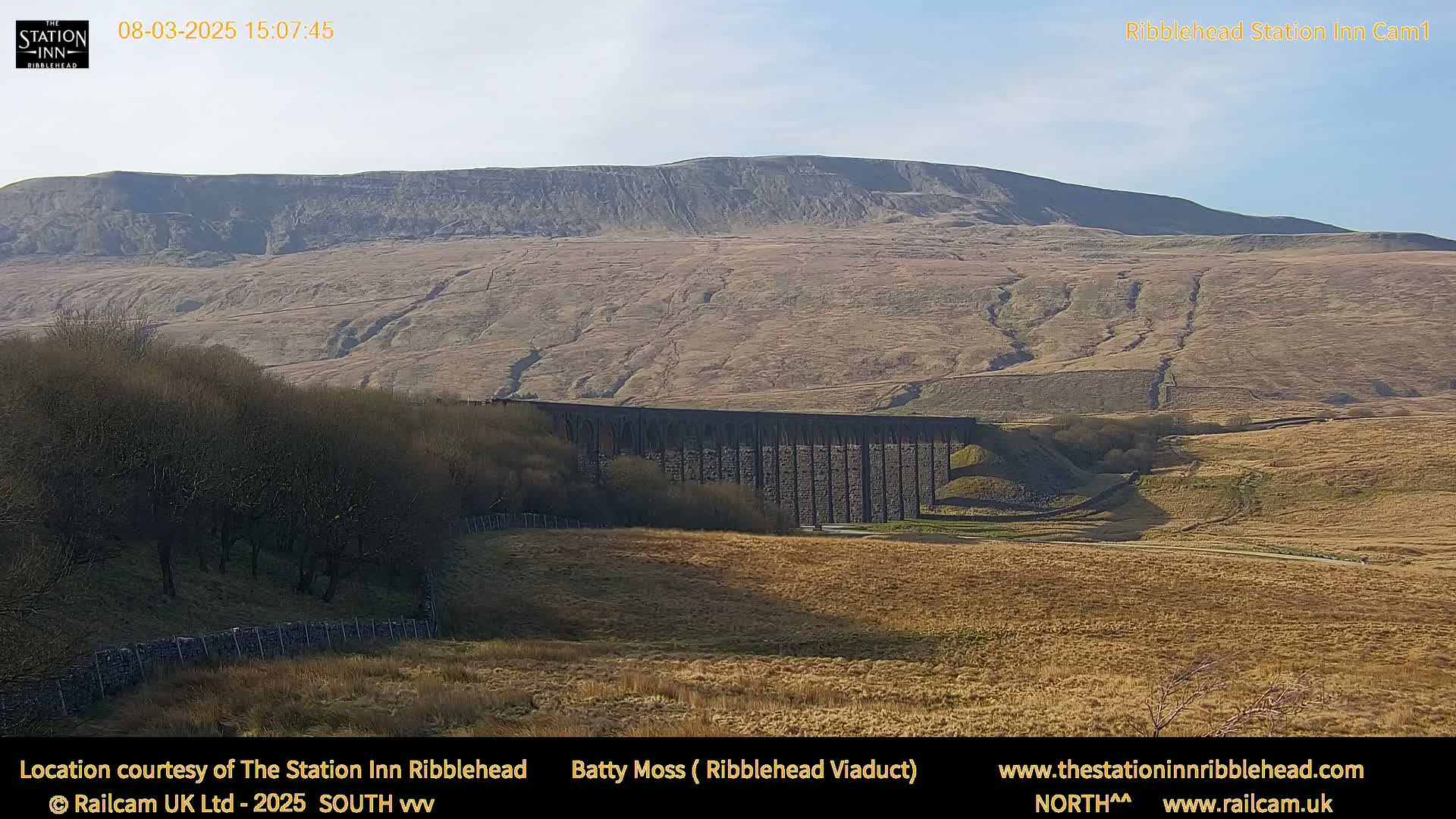 A long stone viaduct stretches across a brown, hilly landscape under a clear, bright sky.