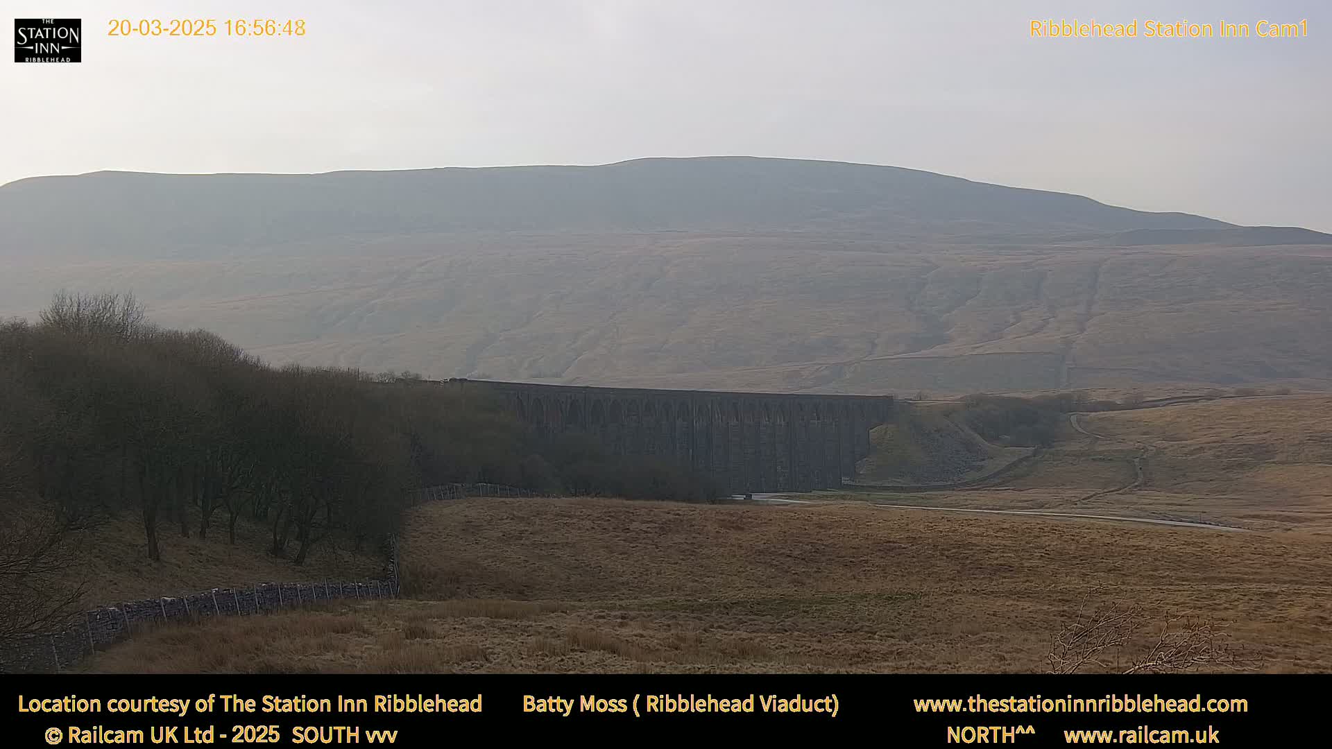A long stone viaduct stretches across a valley under a hazy sky, with a distant, rolling hill in the background.