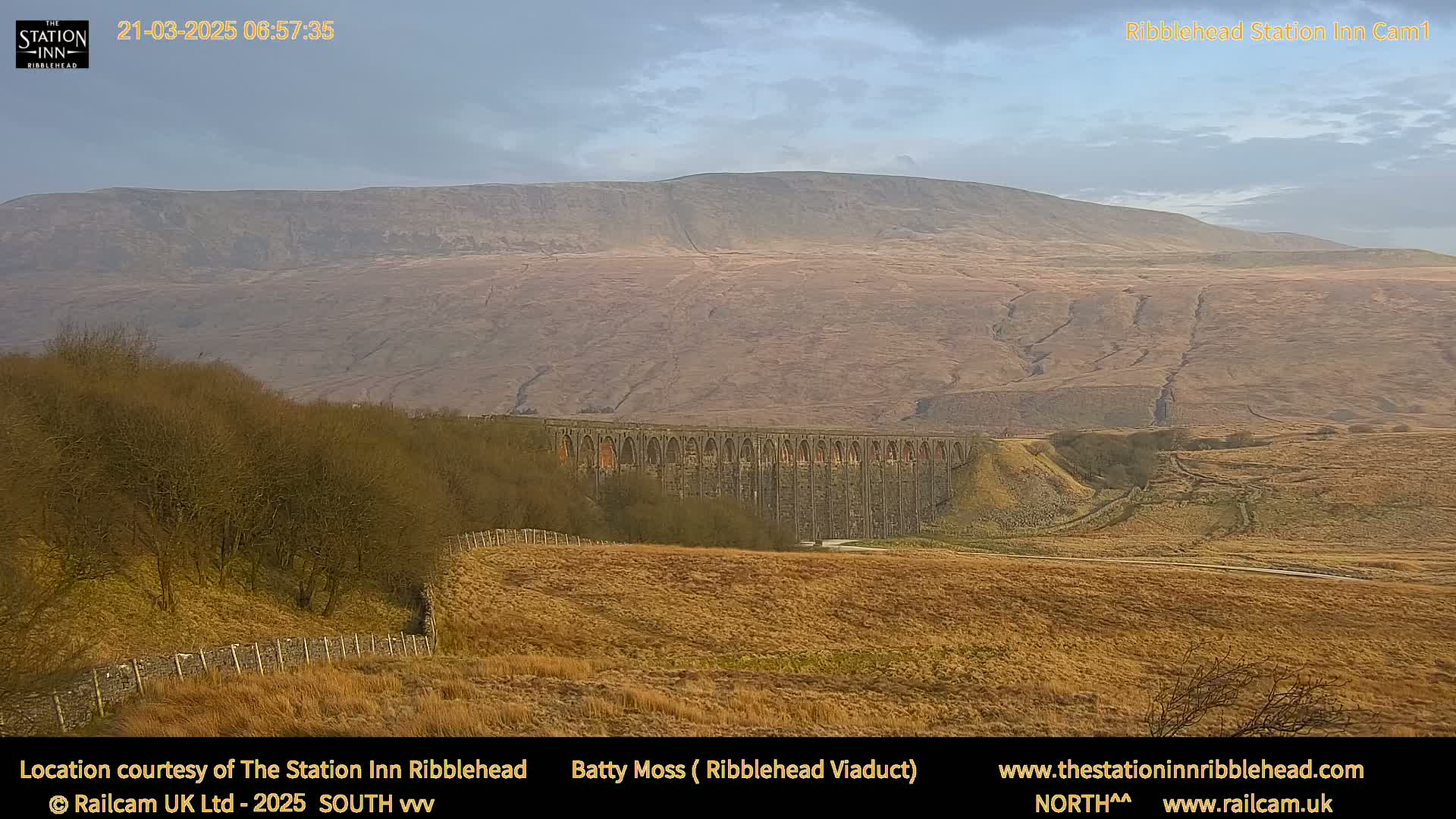 A long stone viaduct stretches across a valley under a mostly cloudy sky, with a large hill rising in the background.