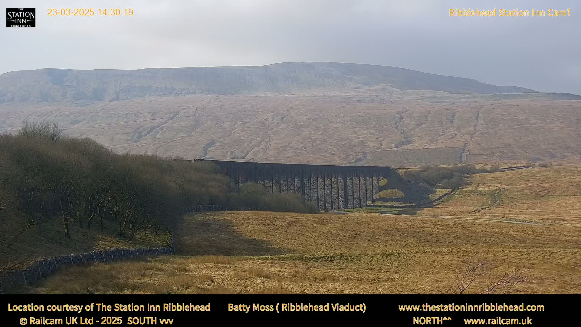 A long stone viaduct sits in a valley below a large, barren hill under a partly cloudy sky.
