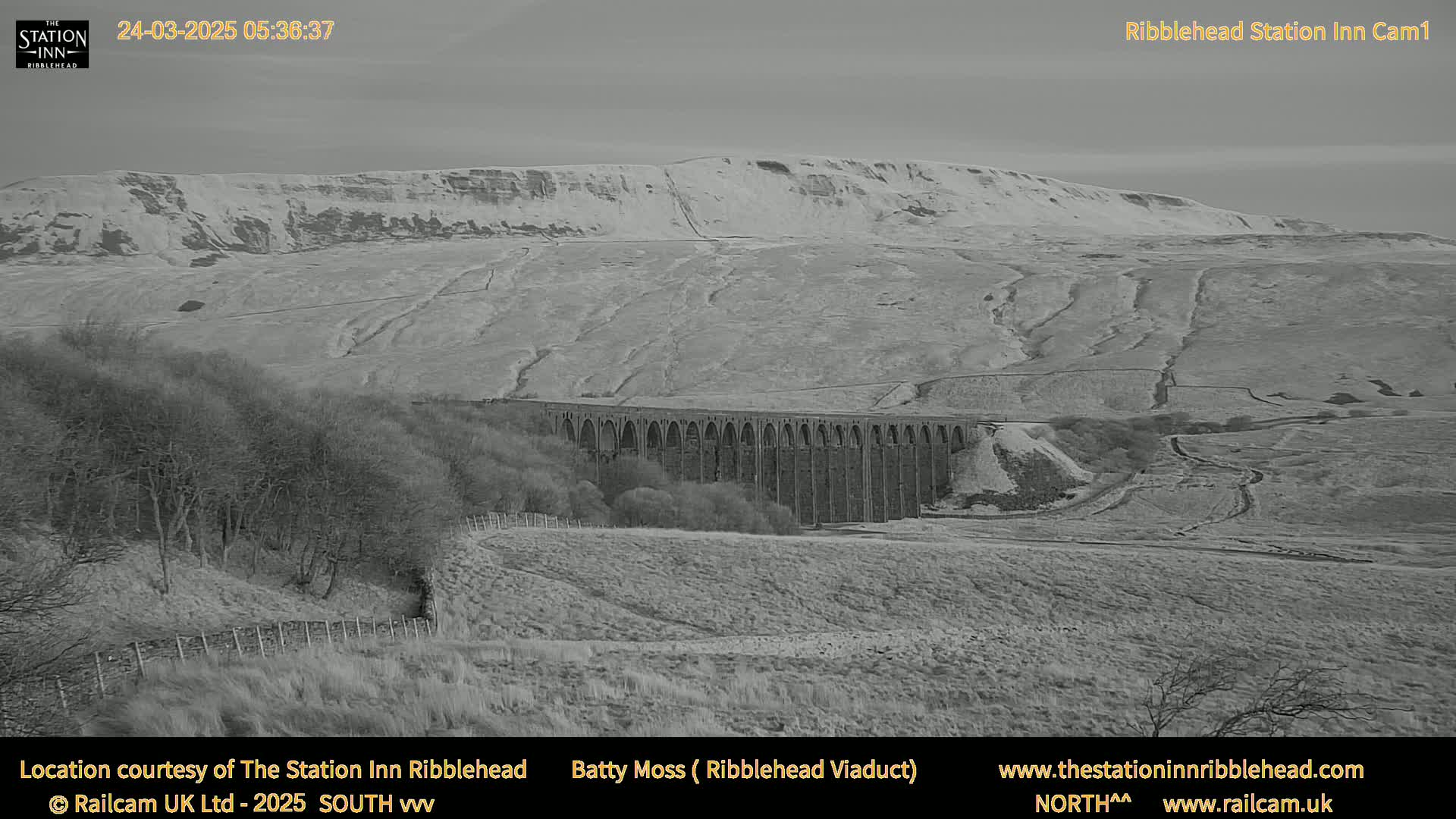 A long stone viaduct stretches across a valley, with a snow-dusted hillside in the background under a mostly clear sky.