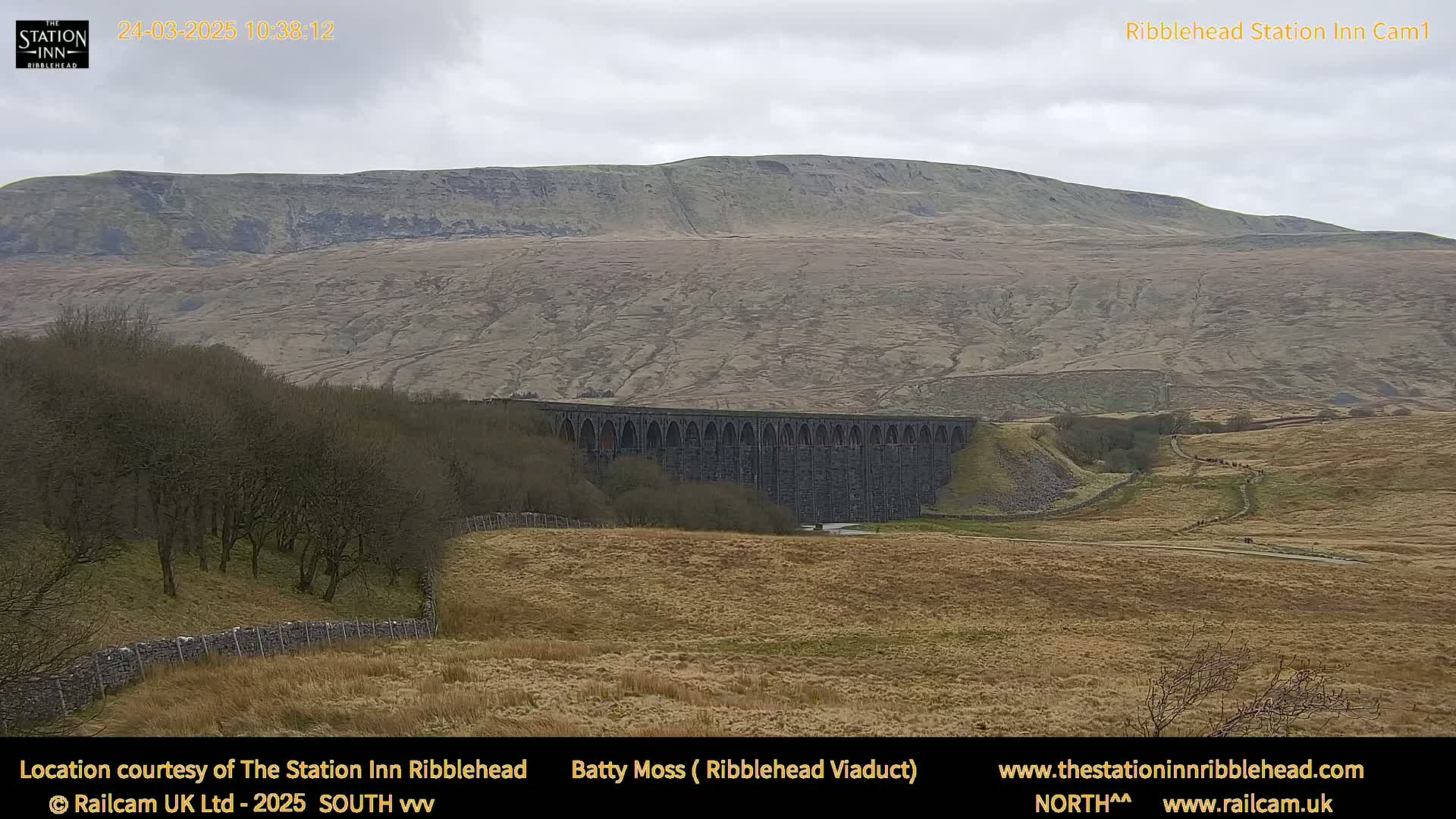 A large stone viaduct sits in a valley beneath a gently sloping, barren hill under an overcast sky.