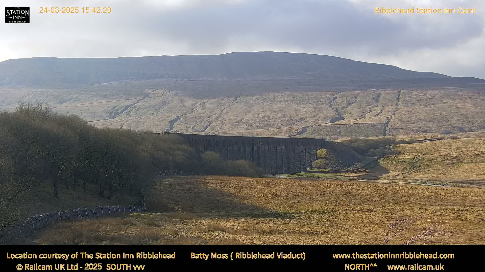 A long stone viaduct sits in a brown, grassy valley beneath a large, pale hill under a mostly cloudy sky.