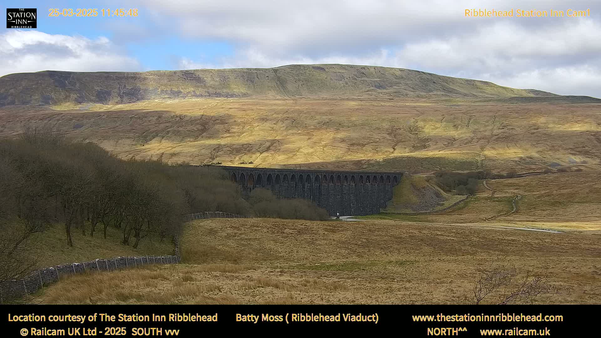 A long stone viaduct sits in a valley below a large, barren hill under a partly cloudy sky.