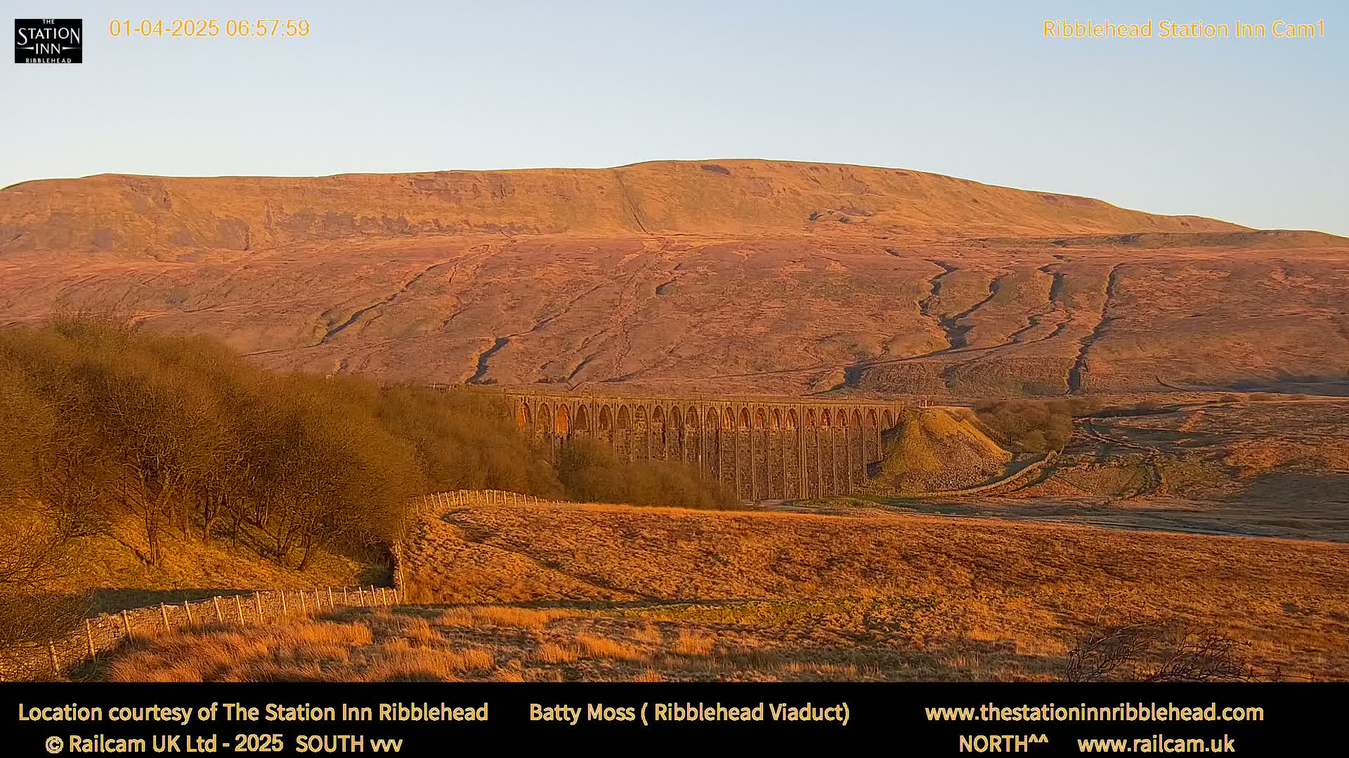 A long stone viaduct sits in a valley beneath a large, golden-brown hill under a clear, light-blue sky.