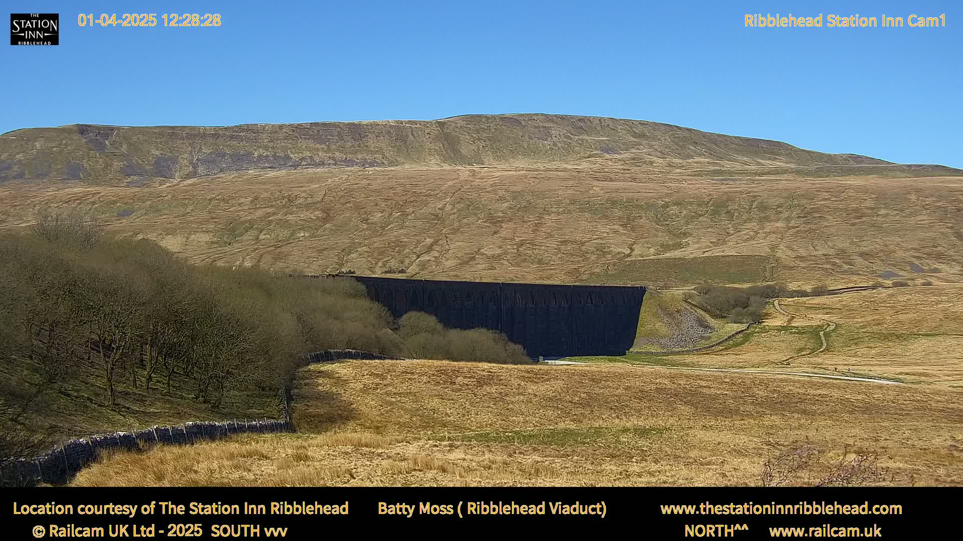 A long, dark viaduct sits in a valley beneath a large, pale brown hill under a clear blue sky.