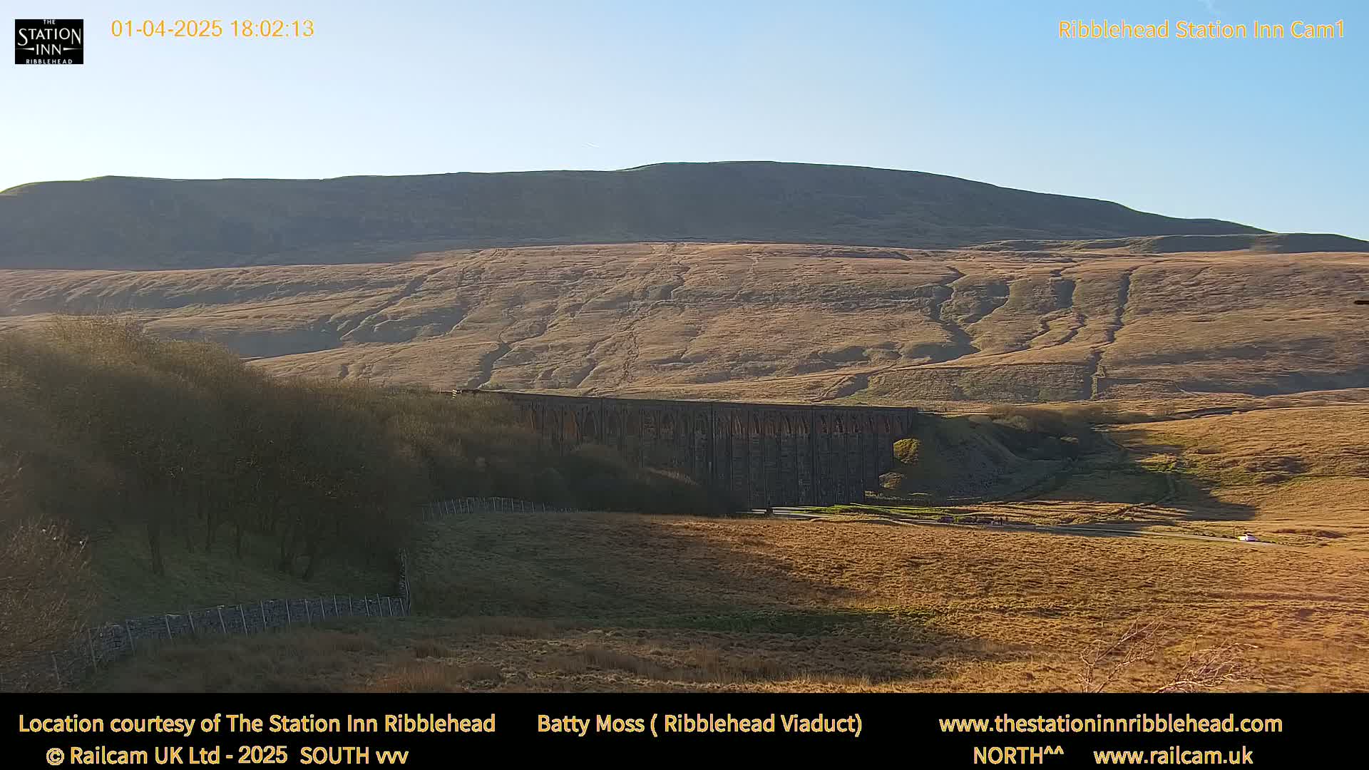 A long stone viaduct stretches across a valley beneath a large, barren hill under a clear, bright sky.