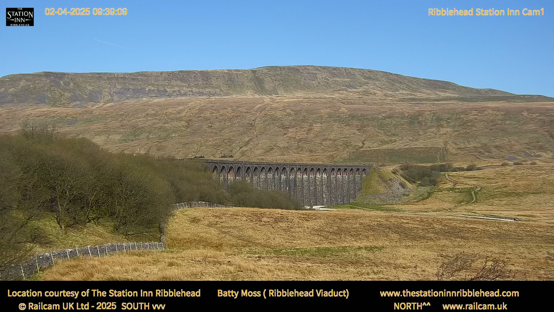 A long stone viaduct sits in a valley below a large, sparsely vegetated hill under a clear blue sky.