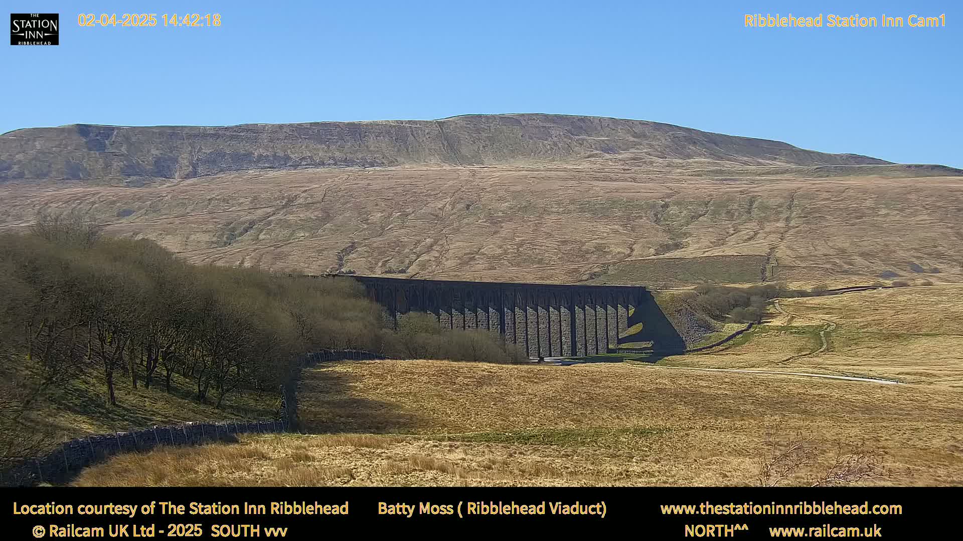 A long stone viaduct sits in a valley below a large, barren hill under a clear blue sky.