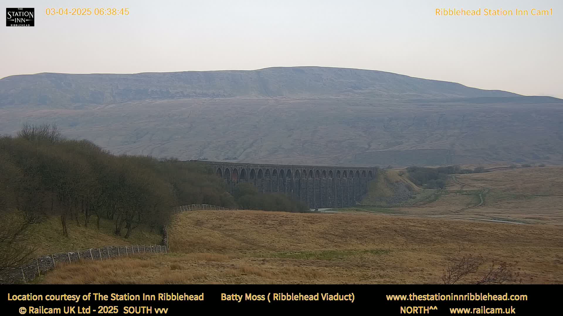 A long stone viaduct sits in a valley below a large, hazy hill under a cloudy sky.