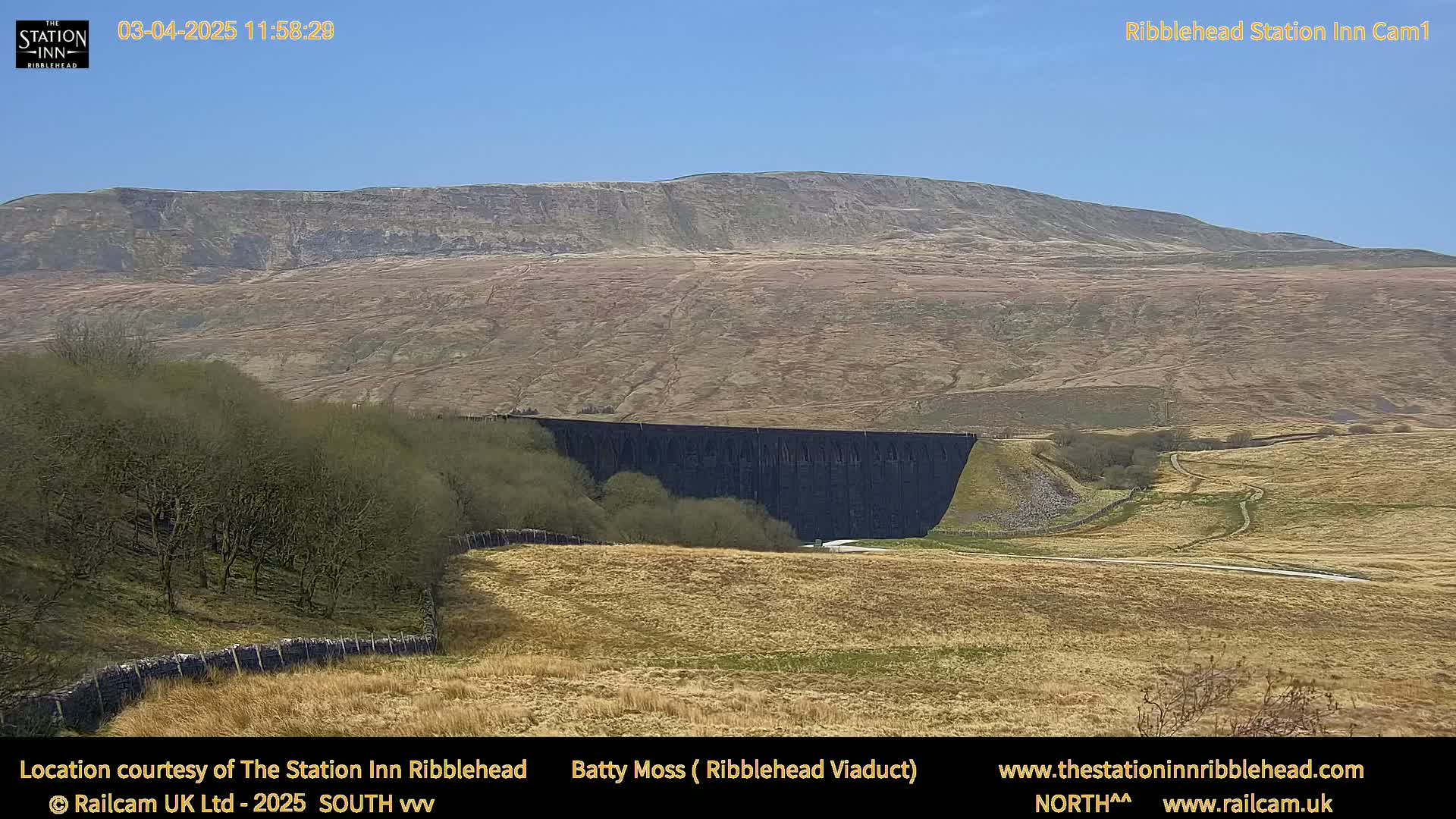 A large, dark viaduct is situated in a valley below a large, light-brown hill under a clear blue sky.