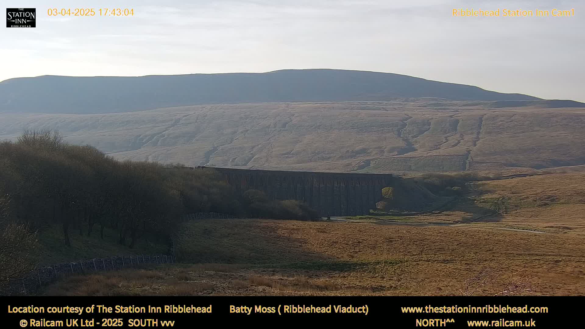 A long stone viaduct sits in a valley beneath a large, gently sloping hill under a clear, bright sky.