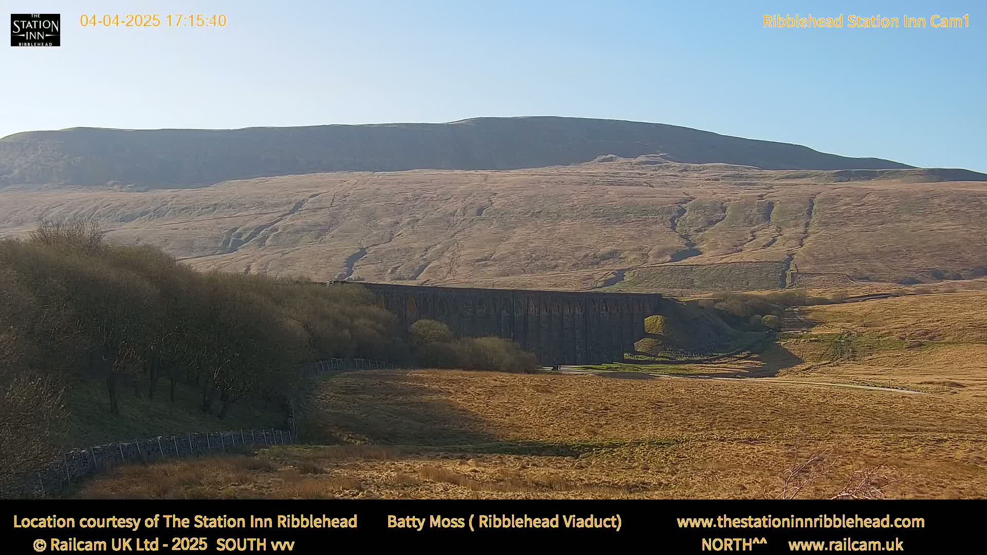 A long stone viaduct sits in a valley beneath a large, gently sloping hill on a sunny day.