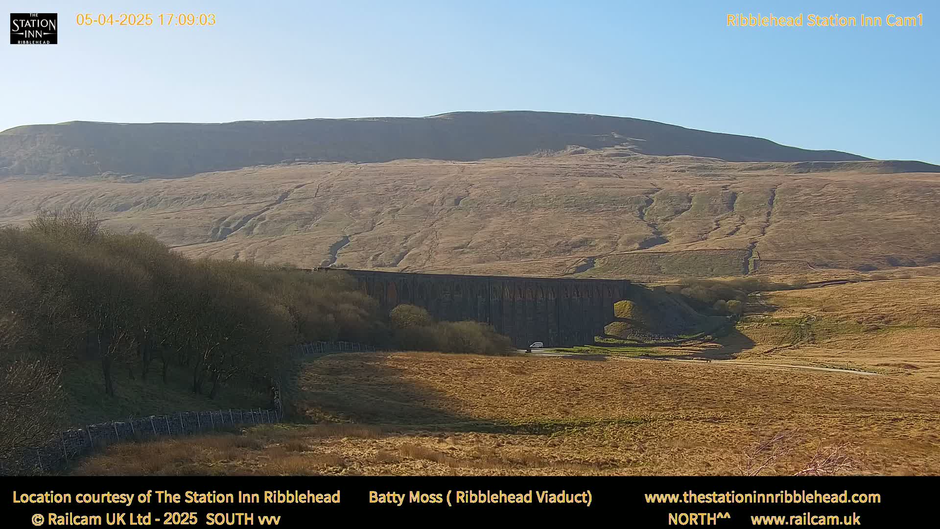 A long stone viaduct sits in a valley beneath a large, tan-colored hill under a clear, sunny sky.