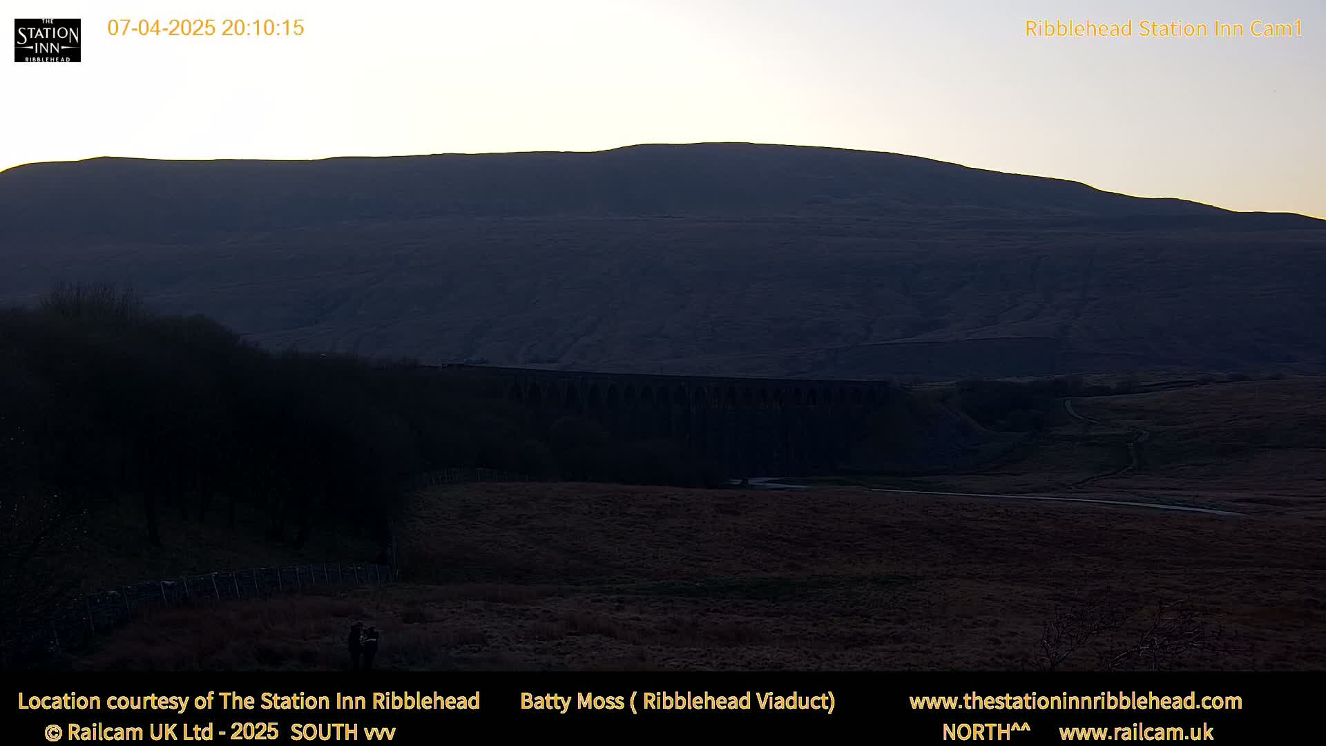 A large, dark hill is visible at dusk, with a long stone viaduct in the foreground.