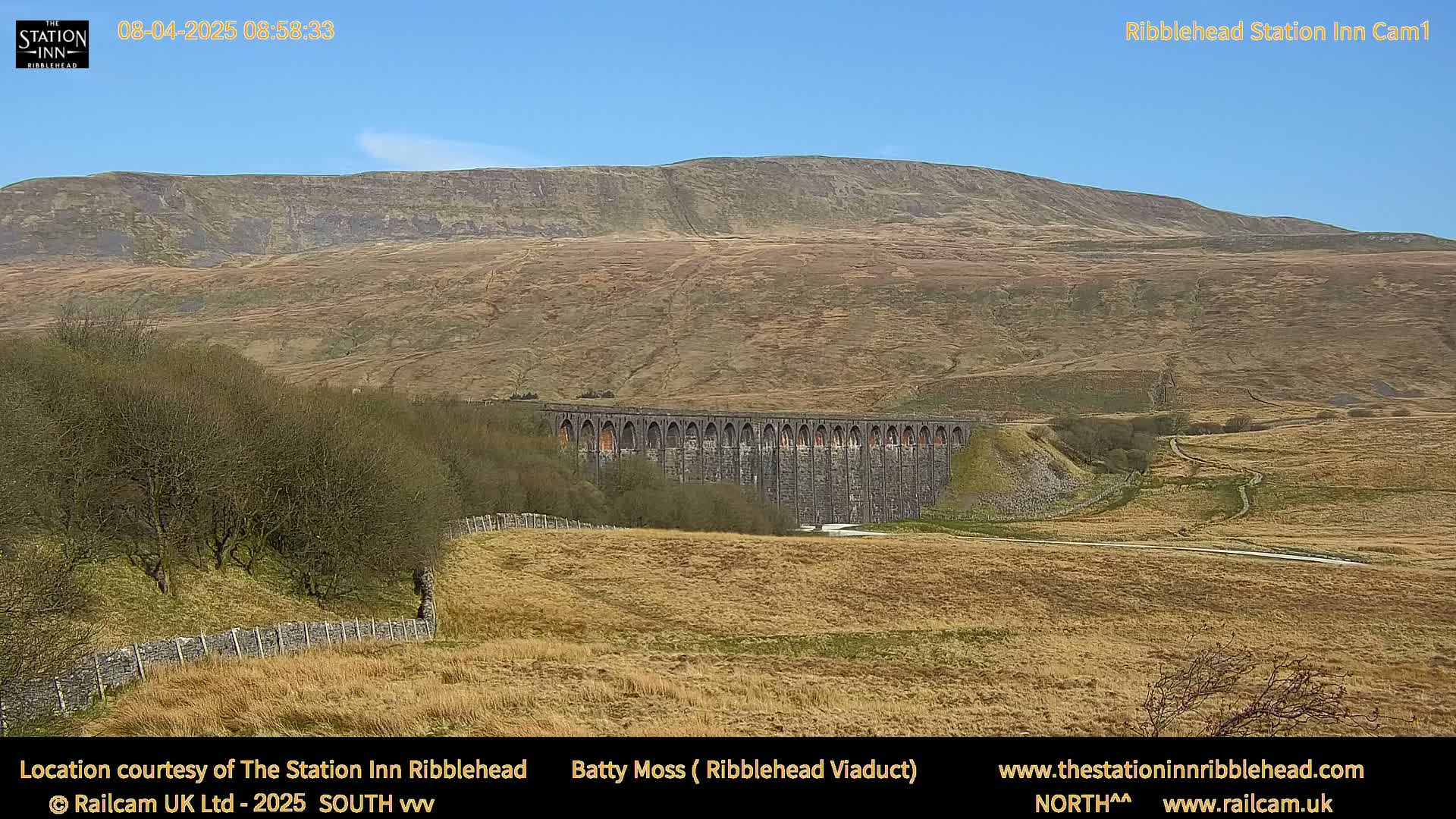 A long stone viaduct sits in a valley under a clear blue sky, with a large, flat-topped hill in the background and dry, brown-yellow fields surrounding it.