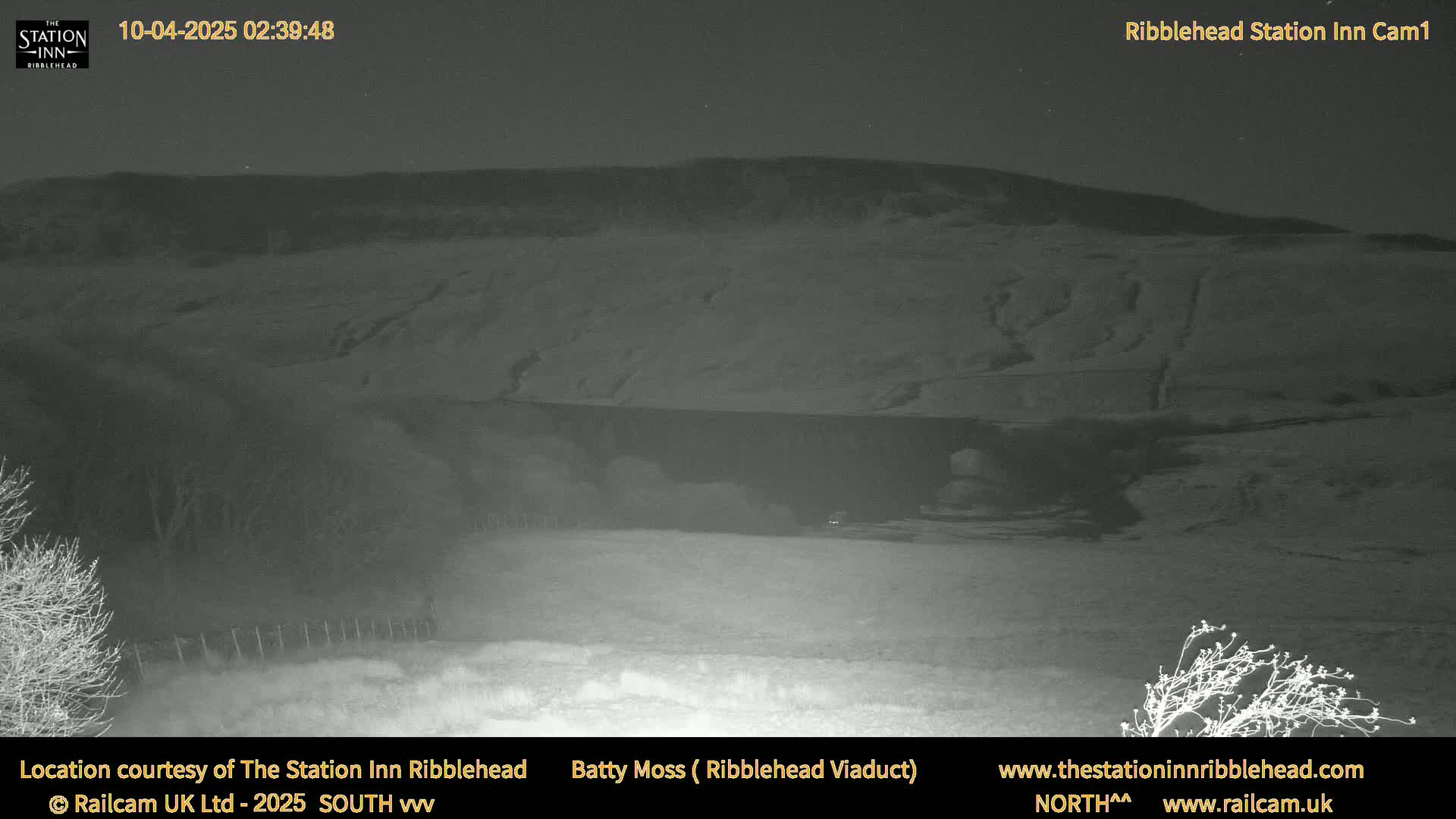 A nighttime, low-light view of a dark, flat landscape with a hill in the background under a clear sky.