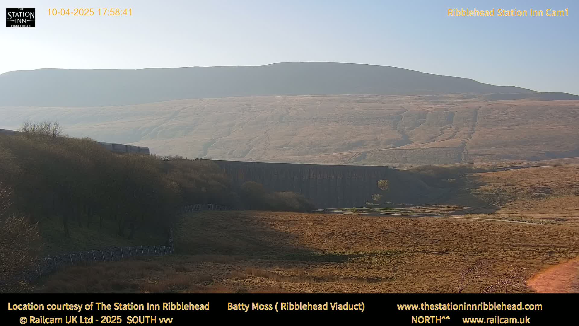 A long stone viaduct stretches across a valley, with a hazy mountain in the background under a clear sky.