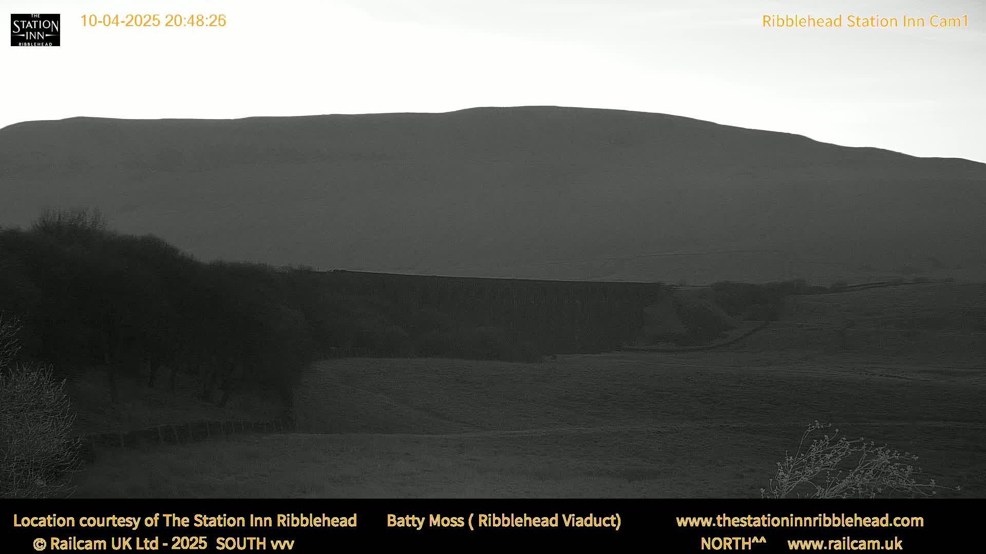 A dark, low-light view of a hill and a long, low structure, possibly a wall or dam, in a rural landscape.