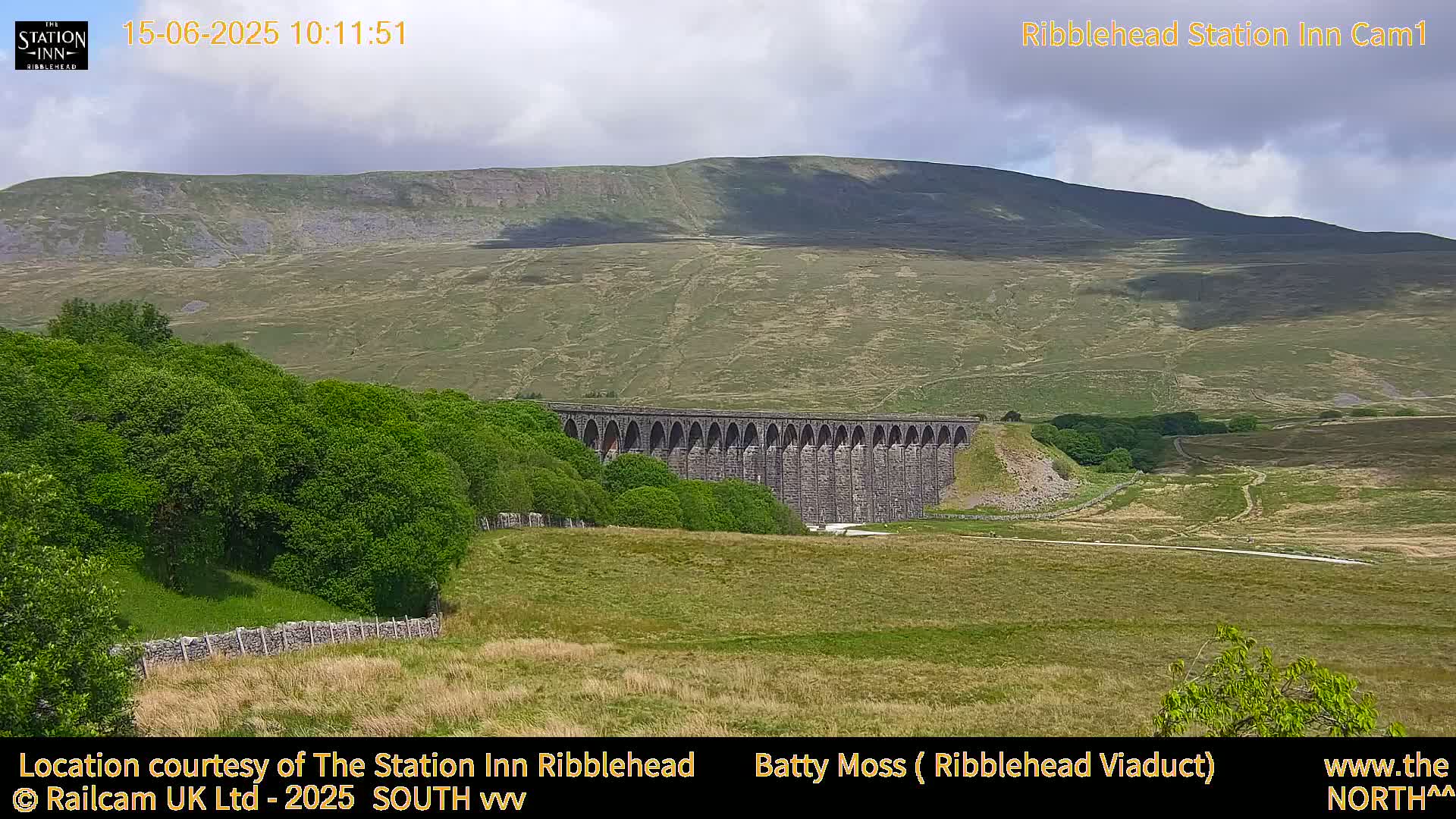 A long stone viaduct is nestled in a valley beneath a gently sloping, light-brown hill under a partly cloudy sky.