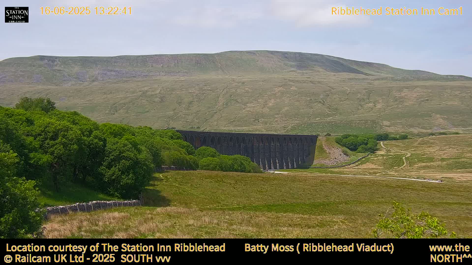 A large stone viaduct sits in a valley beneath a gently sloping, brown hill under a mostly clear sky.