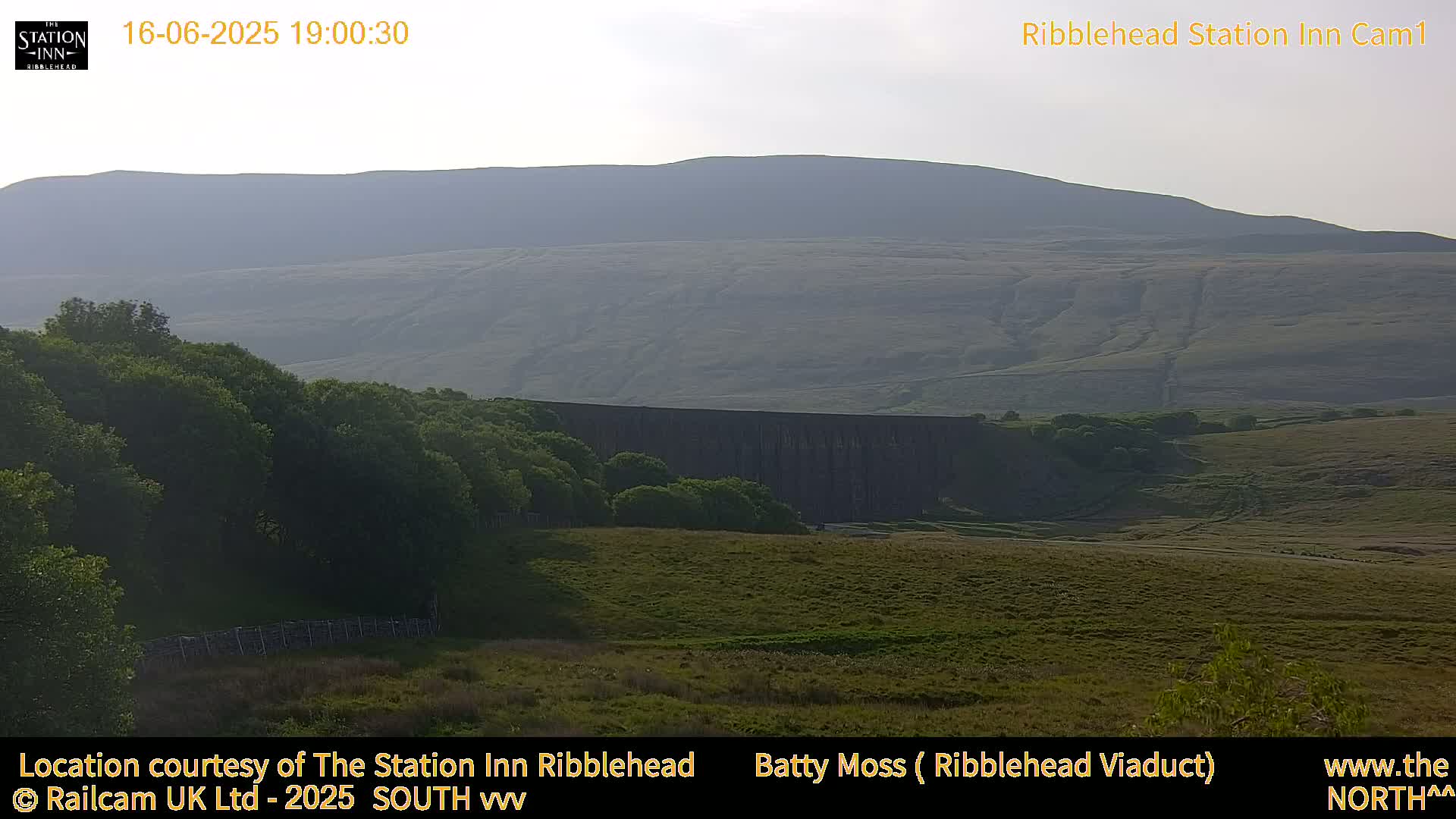 A long, dark viaduct is nestled amongst green hills and trees under a clear, light evening sky.