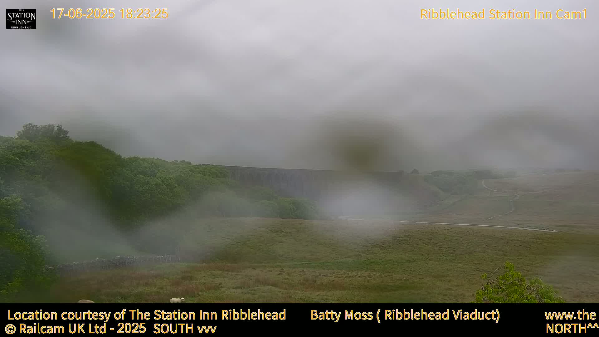 A misty view shows a long stone structure in a valley surrounded by green hills and a few sheep in a field.