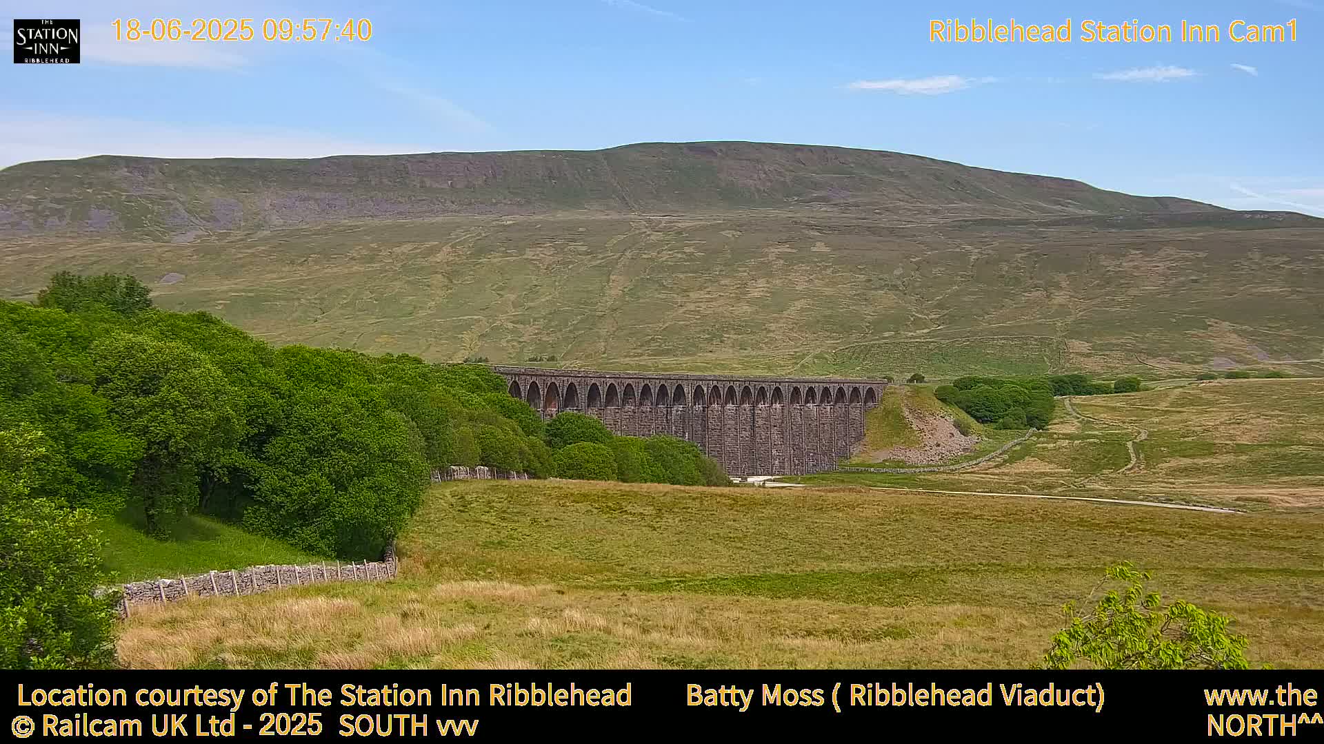 A long stone viaduct sits in a verdant valley under a clear blue sky, with a large hill rising in the background.
