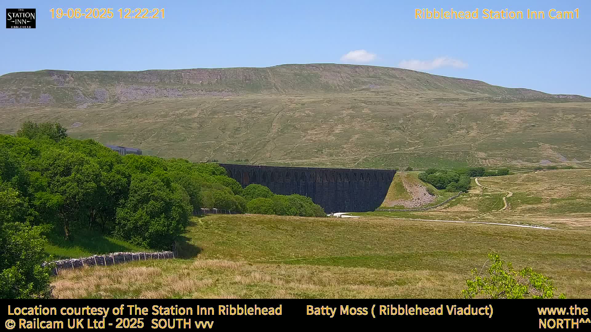 A large stone viaduct sits in a grassy valley beneath a large, gently sloping hill under a clear blue sky.