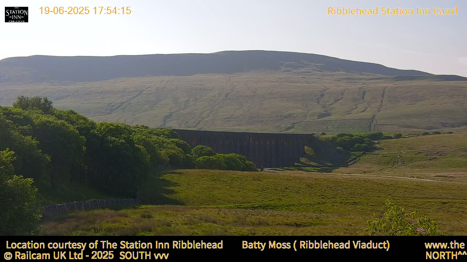 A long stone viaduct sits in a valley beneath a large, gently sloping hill under a clear, bright sky.