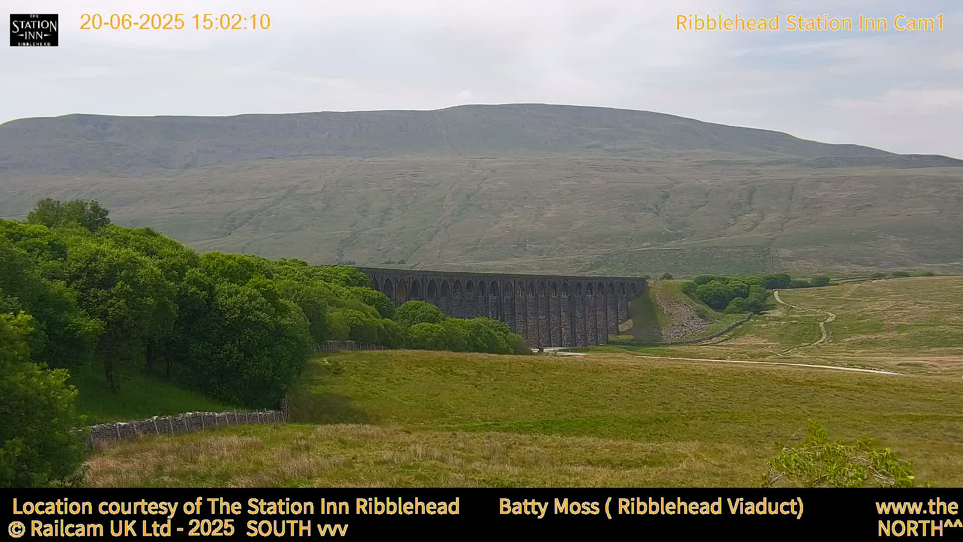 A long stone viaduct is nestled in a green valley beneath a large, gently sloping hill under an overcast sky.