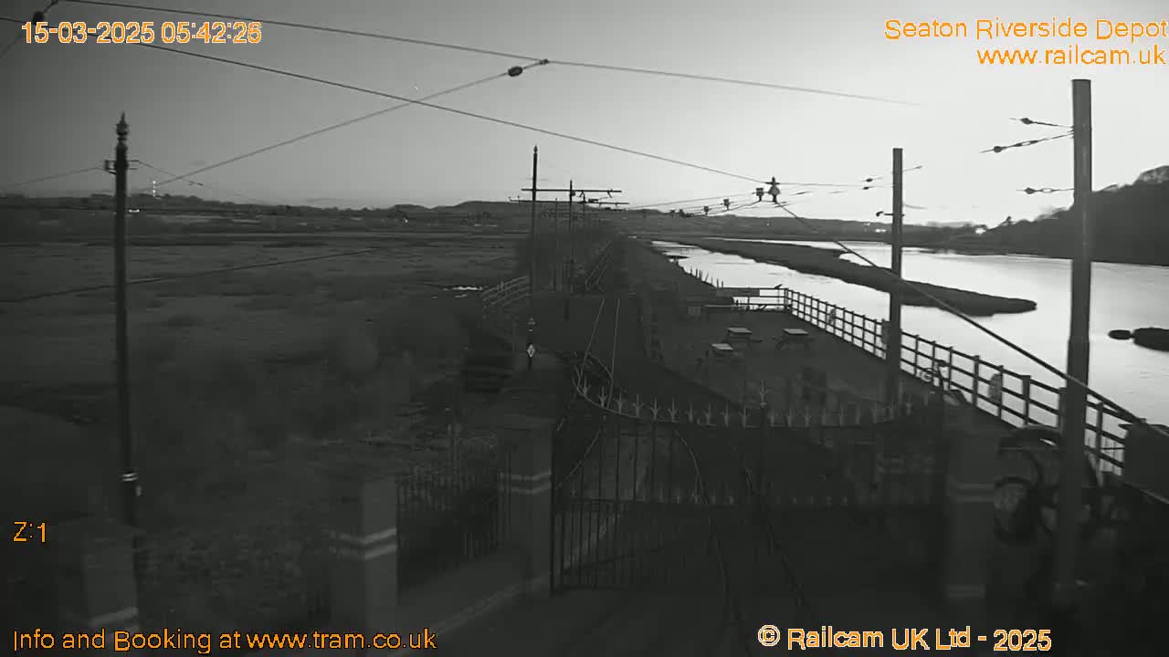 A black and white, twilight view of a riverside depot with overhead power lines, a calm body of water, and a gated entrance.