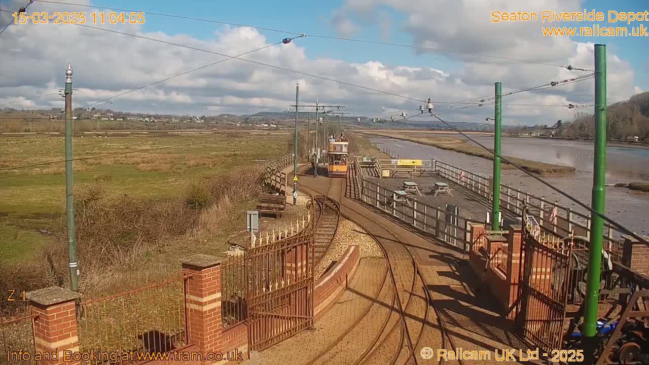 A yellow tram approaches a depot on a sunny day with partly cloudy skies, alongside a body of water and marshland.