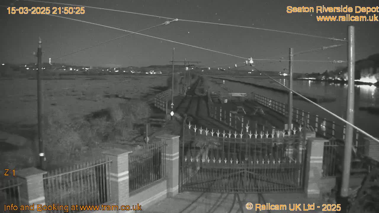 A nighttime, black and white view of a gated area near a body of water, with overhead wires and distant lights visible under a clear sky.