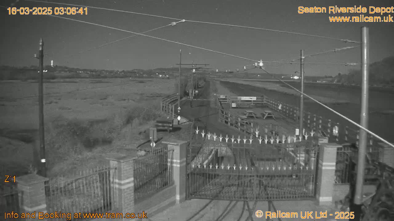 A nighttime, black-and-white view of a gated entrance to a depot, with overhead wires and a flat, open area beyond.