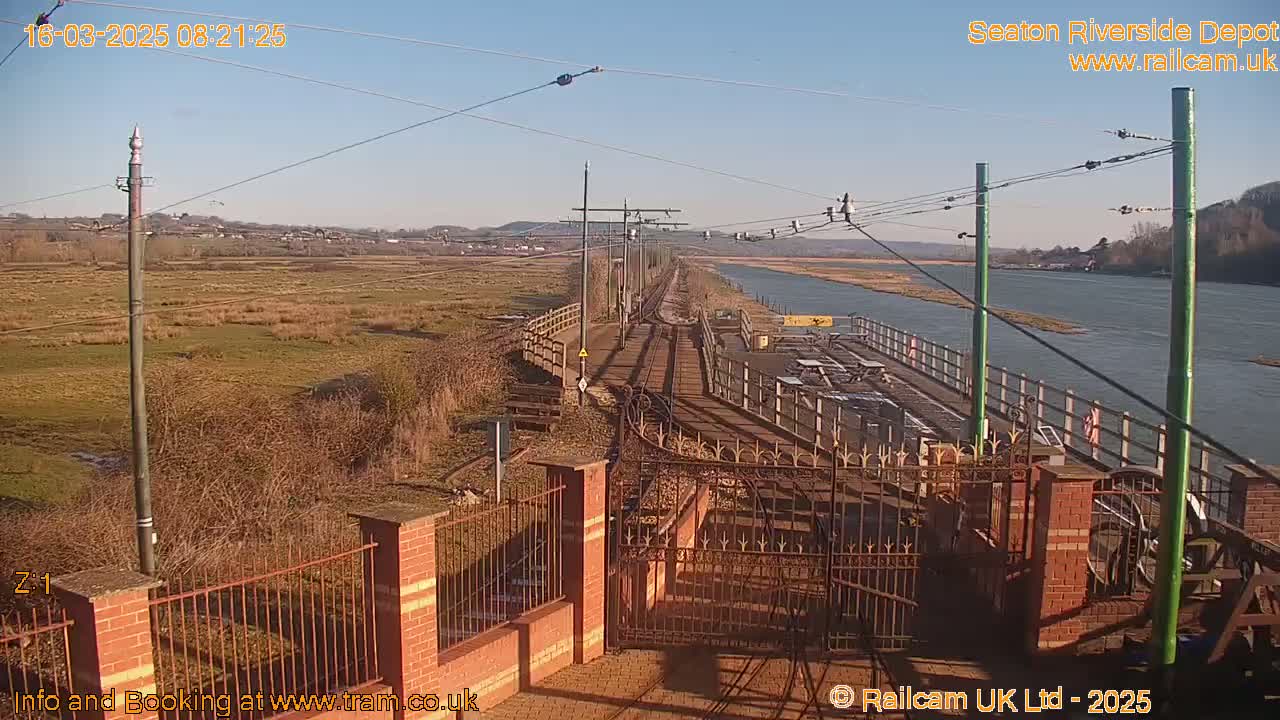 A sunny day reveals a train depot situated beside a river, with a rail line extending into the distance across marshland.