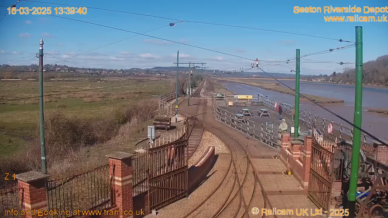 A sunny day at a train depot near a body of water, showing train tracks and a fenced-in area with picnic tables.