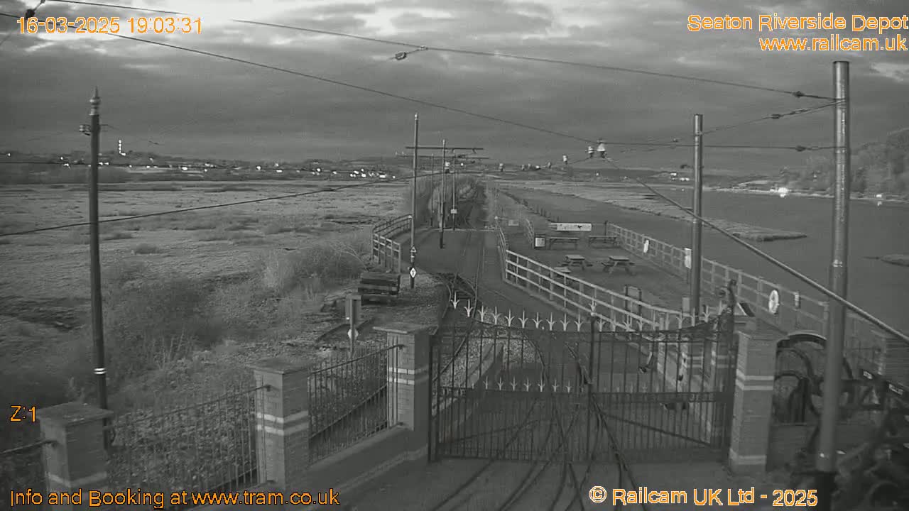 A grayscale image shows an overcast evening view of a railway depot with a gated entrance, fences, and a paved area leading to a body of water under a cloudy sky.